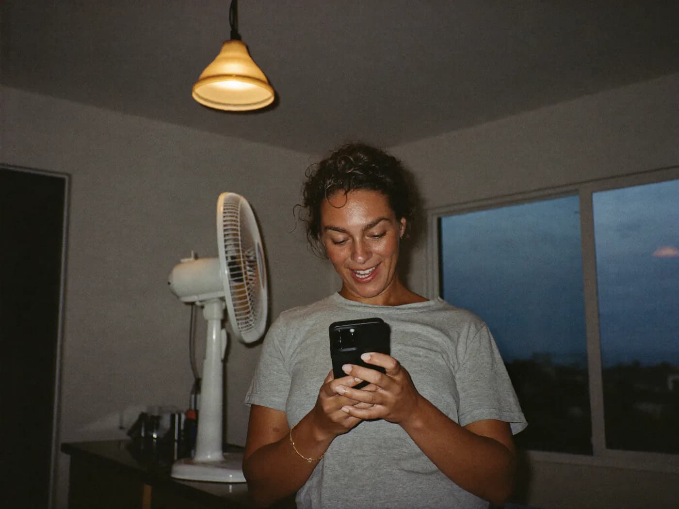 A homeowner smiles at a text message in her kitchen on a hot evening, a fan running behind her.